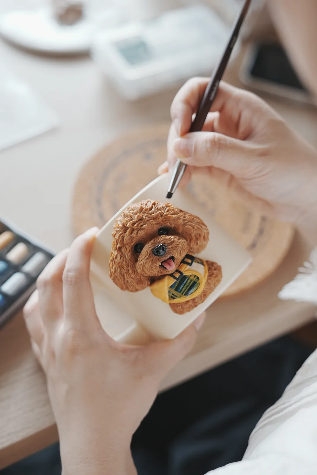 筆を持った手で、チェック柄のバンダナをつけた茶色のトイプードルのマグカップに色を入れているところ