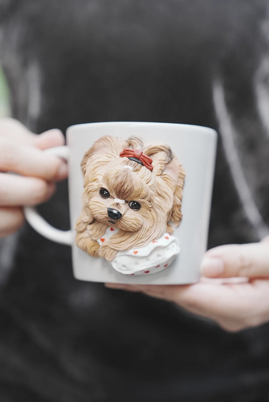リボンを結びハート柄の服を着た長毛の犬を立体にした白いマグカップを、両手で持つところ